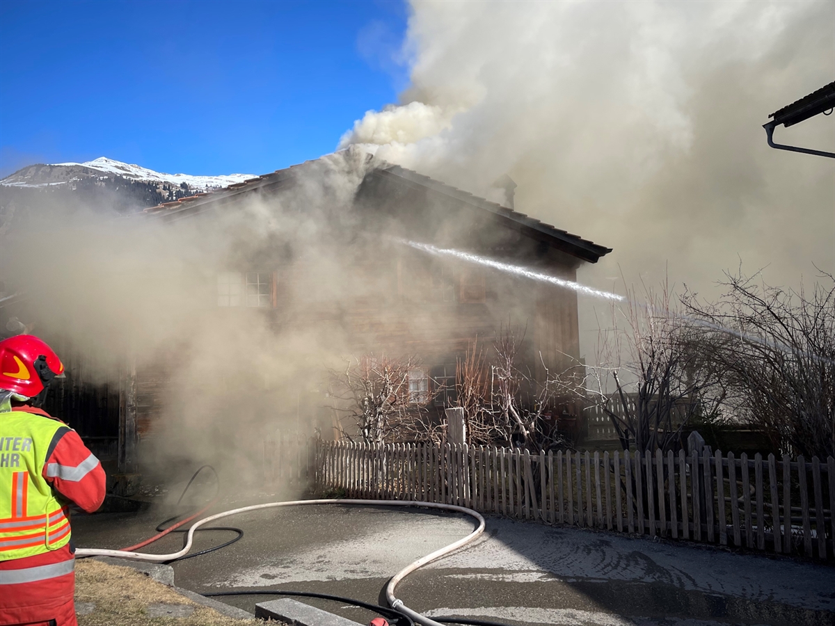 Der blaue Himmel wird mit dunklem Rauch verzerrt, welcher vom brennenden Gebäude stammt. Der Einsatzleiter Feuerwehr überwacht die Löscharbeiten seiner Einsatzkräfte.