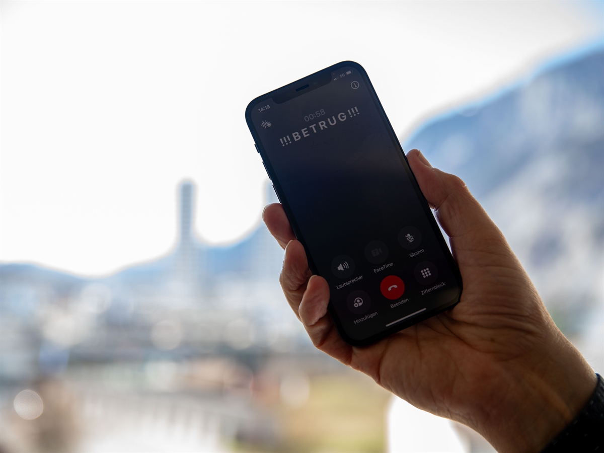 Verschwommene Landschaft im Hintergrund. Im Vordergrund eine Hand, welche ein Mobiltelefon hält. Auf diesem der Schriftzug !!! Betrug !!!