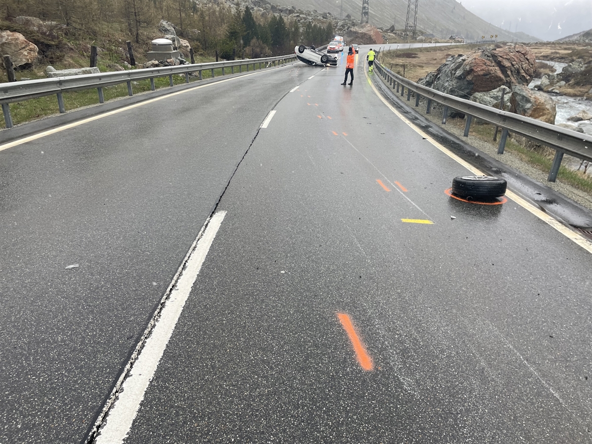 Im Vordergrund das abgerissene Rad. In Entfernung ein Polizist, das auf dem Dach liegende Auto, ein Polizeiauto und ein Rettungswagen.