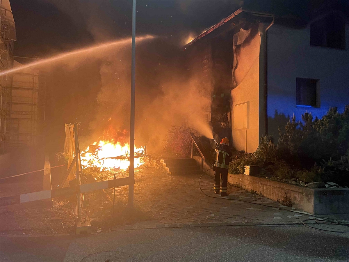 Der Wohnanhänger ist in Flammen. Ein Feuerwehrmann löscht das Feuer. Das stark beschädigte Wohnhaus ist auch erkennbar.