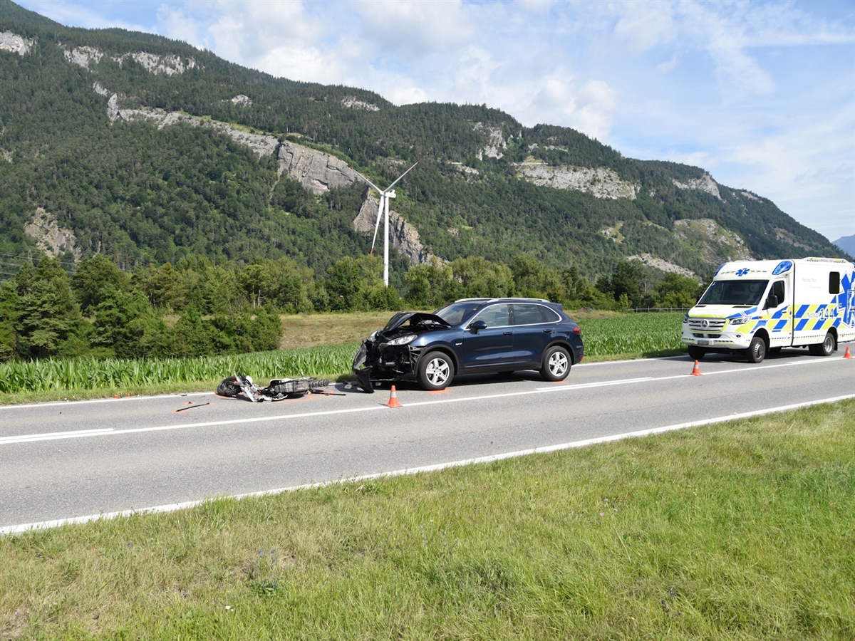 Das in zwei Teile zerbrochene Motorrad liegt vor dem Personenwagen. Dieser ist an der Front erheblich beschädigt. Dahinter ein Rettungswagen. Im Hintergrund Berge, die den Horizont abgrenzen. 