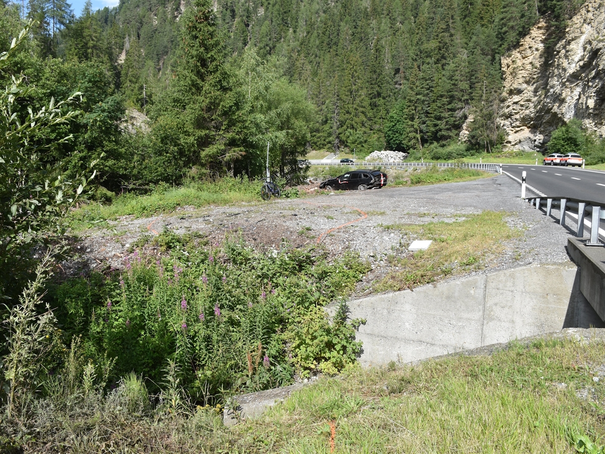 Tagaufnahme mit dem Grünstreifen und dem Bachbett. Das total beschädigte Auto in der Endlage.