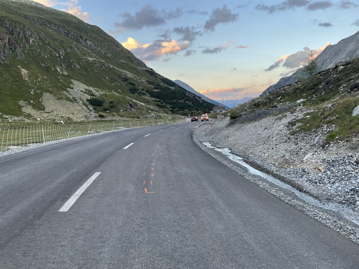 Die Rechtskurve mit auf der Strasse eingezeichneten Unfallspuren. In Entfernung ein Polizeiauto. Blick in die Ferne mit Bergen die den Horizont abgrenzen.