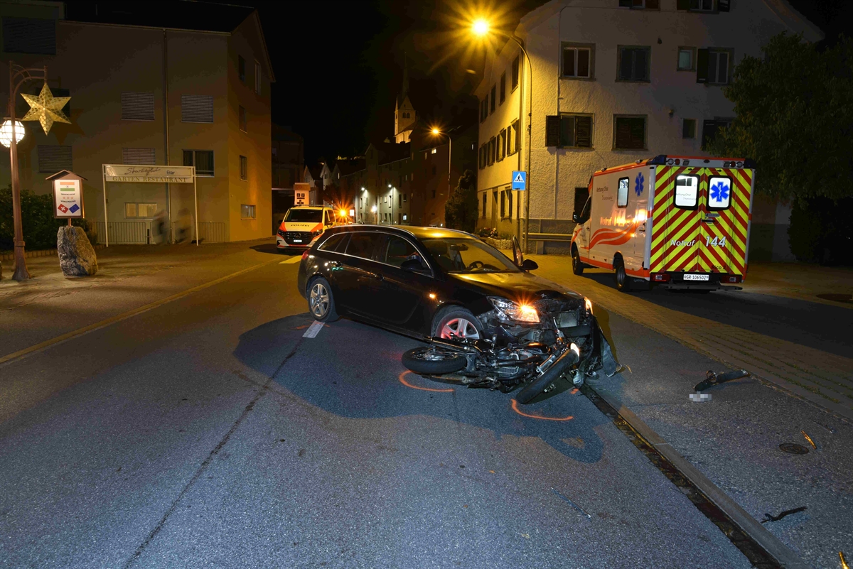 Es ist dunkel. Der an der Front beschädigte schwarze beteiligte Personenwagen steht schräg mitten auf der Strasse. Das schwarze Motorrad liegt beschädigt vor der Front des Autos am Boden. Rechts auf dem Parkplatz steht die Ambulanz. Im Hintergrund steht ein Polizeifahrzeug.