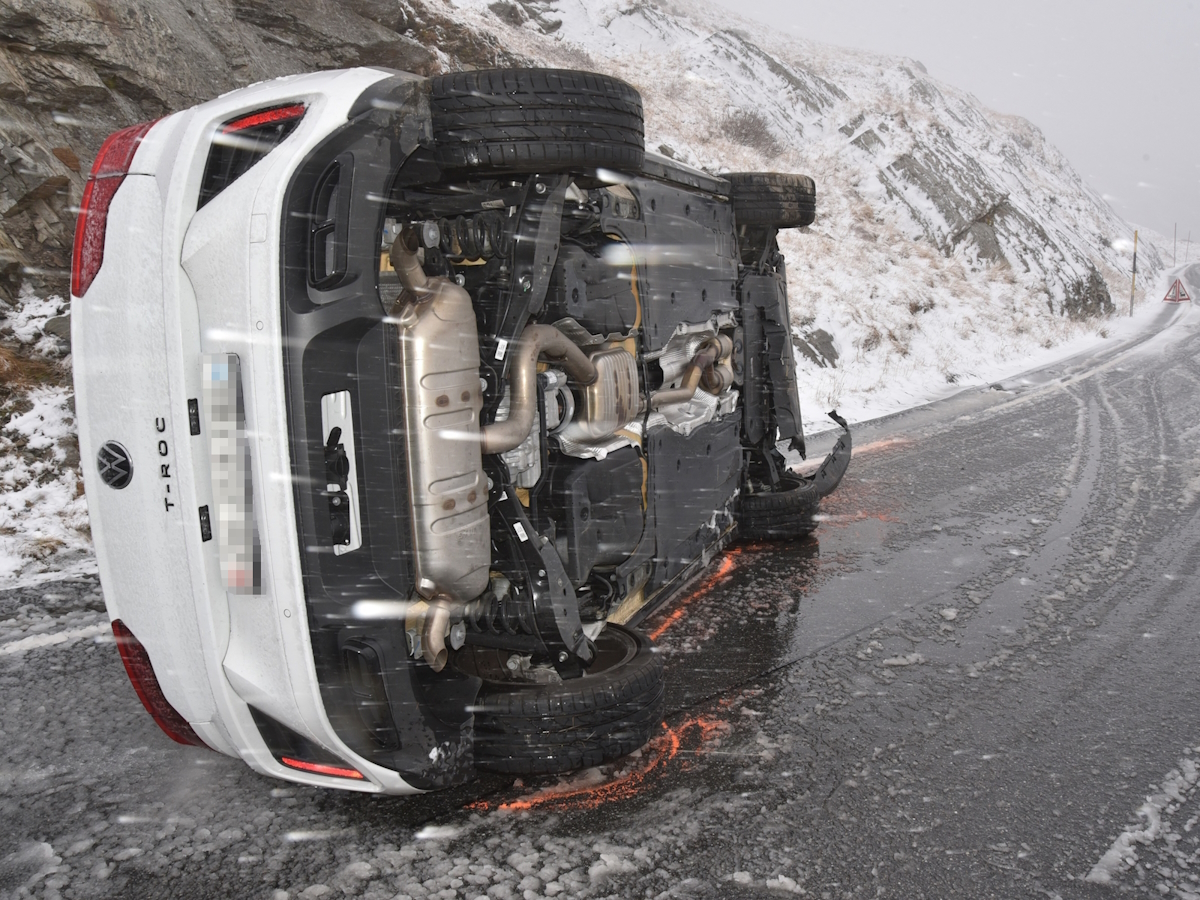 Das auf der Seite liegende Auto mit Sommerreifen auf der schneebedeckten Splügenpass-Strasse. Im Hintergrund die frisch verschneite Landschaft