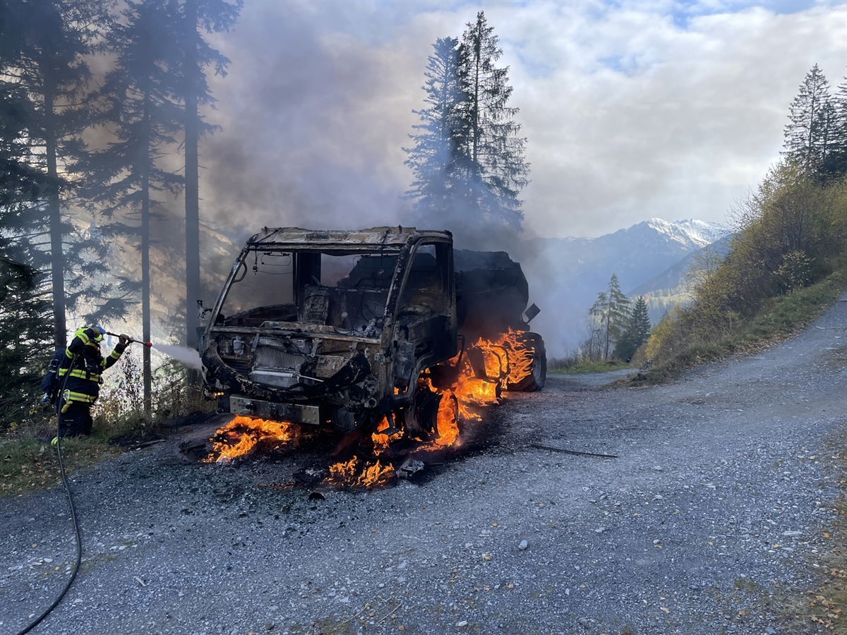 Ein Feuerwehrmann beim Löschen des brennenden Motorkarren. Unter diesem lodern Flammen hervor. Starker Rauch trübt die Sicht auf die umliegenden winterlichen Berge