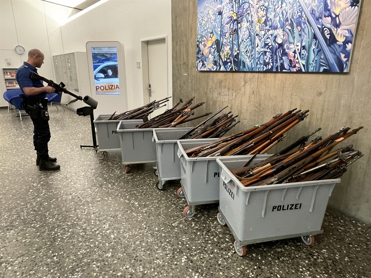 Ein Polizist entlädt eine Waffe im Zwischengang des Polizeikommandos in Chur. 5 grosse Plastikbehälter stehen in Reih und Glied, gefüllt mit Langwaffen.