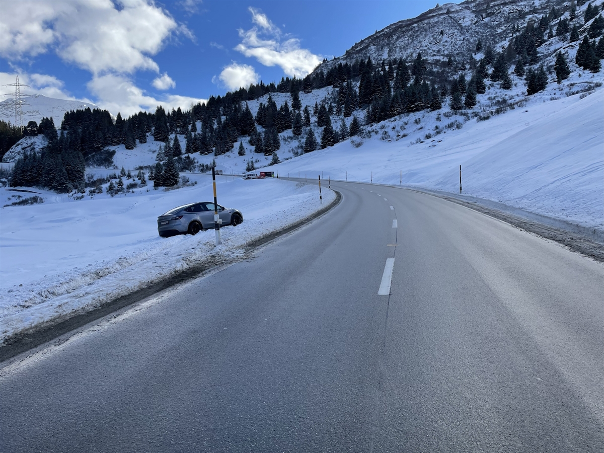 Winterlandschaft. Links neben der Strasse ein Auto im Schnee. Der zumeist heitere Horizont wird durch Gebirgslandschaft abgegrenzt.