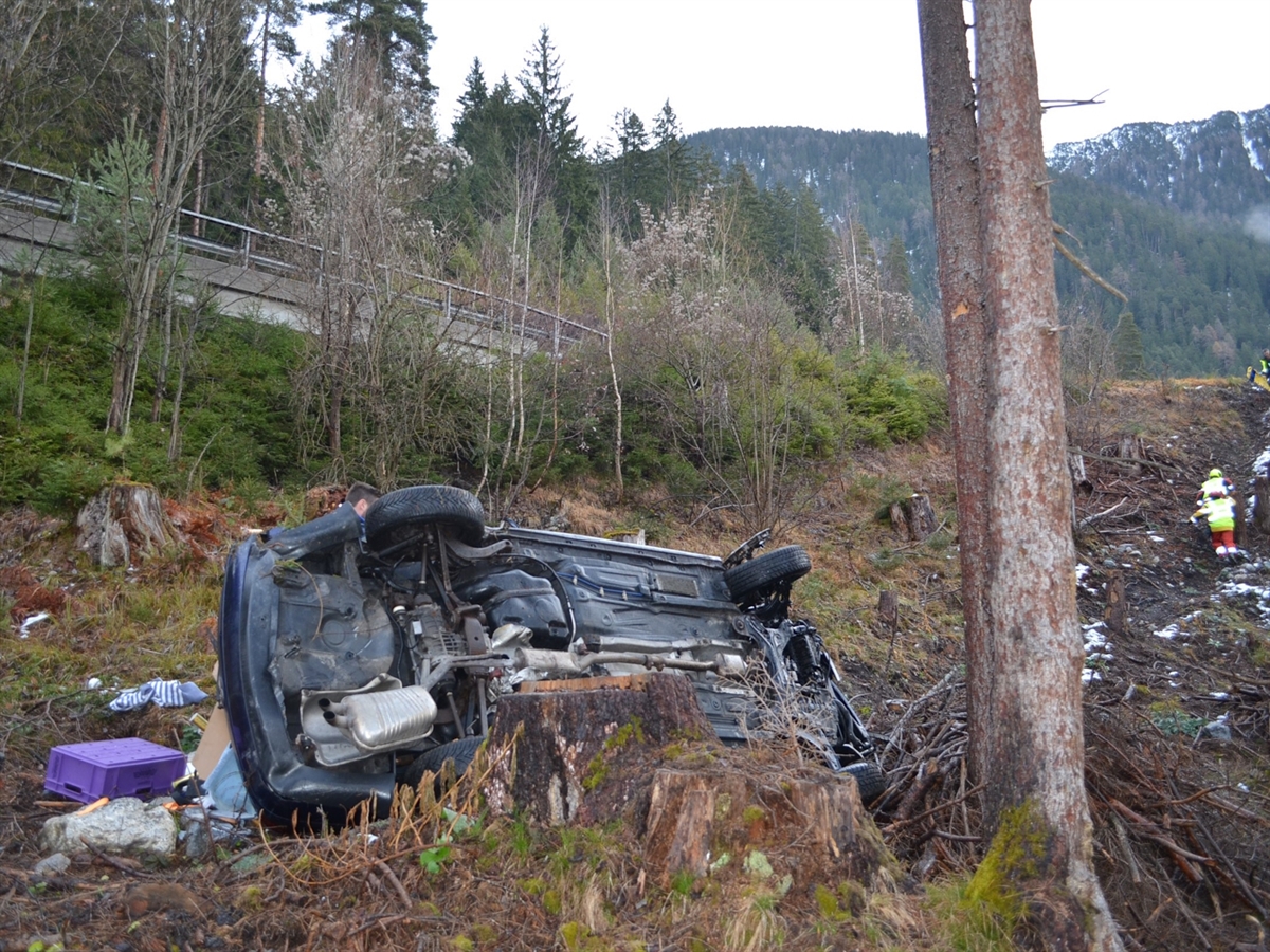 Das Auto liegt auf der linken Seite an einem Wurzelstock angelehnt. Viel mitgeführtes Material liegt ausserhalb des Autos. Feuerwehrleute begeben sich zum Unfallauto. Weiter ist in der oberen Bildhälfte die Leitplanke und Stützmauer der N29 zu sehen.