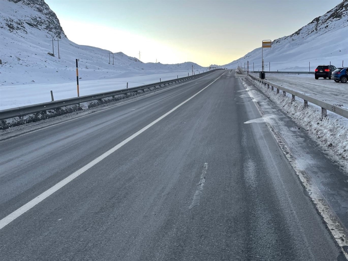 Winterlandschaft. Der Strassenabschnitt in Richtung Passhöhe gesehen. Rechts eine Parkierungsanlage. Berge heben sich vom heiteren Horizont ab.