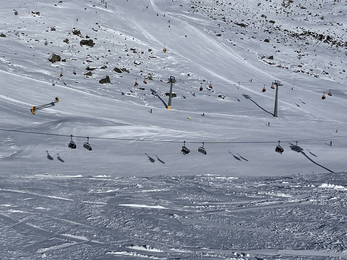 Verschneite Skipiste im Skigebiet mit zwei Sessellift, einzelne Skifahrer fahren über den Hang, im Hintergrund felsdurchsetzte Bergflanken unter klarem Himmel.