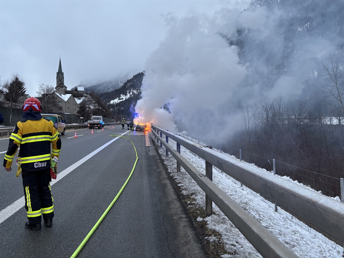 Auf der Autobahn A13 steht der Feuerwehrkommandant und überwacht den Löschvorgang der Feuerwehrleute beim brennenden Auto. Über den Pannenstreifen ist ein Wasserschlauch ausgelegt. Auf der Überholspur fahren Fahrzeuge am Ereignisort vorbei.