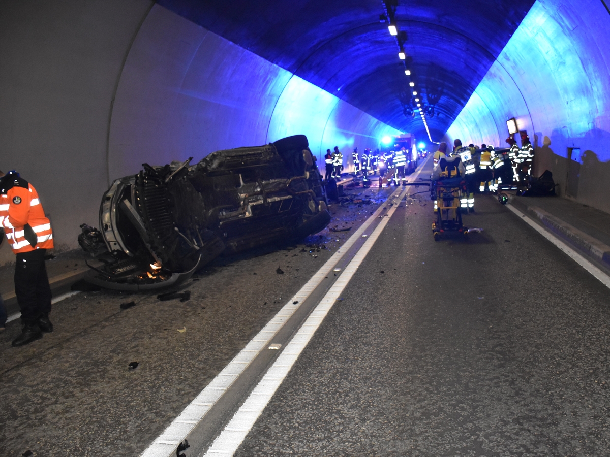 Ein auf der rechten Seite liegendes Auto. In dessen Nahbereich Einsatzkräfte. Blaulicht eines Einsatzfahrzeugs, das den Tunnel illuminiert.