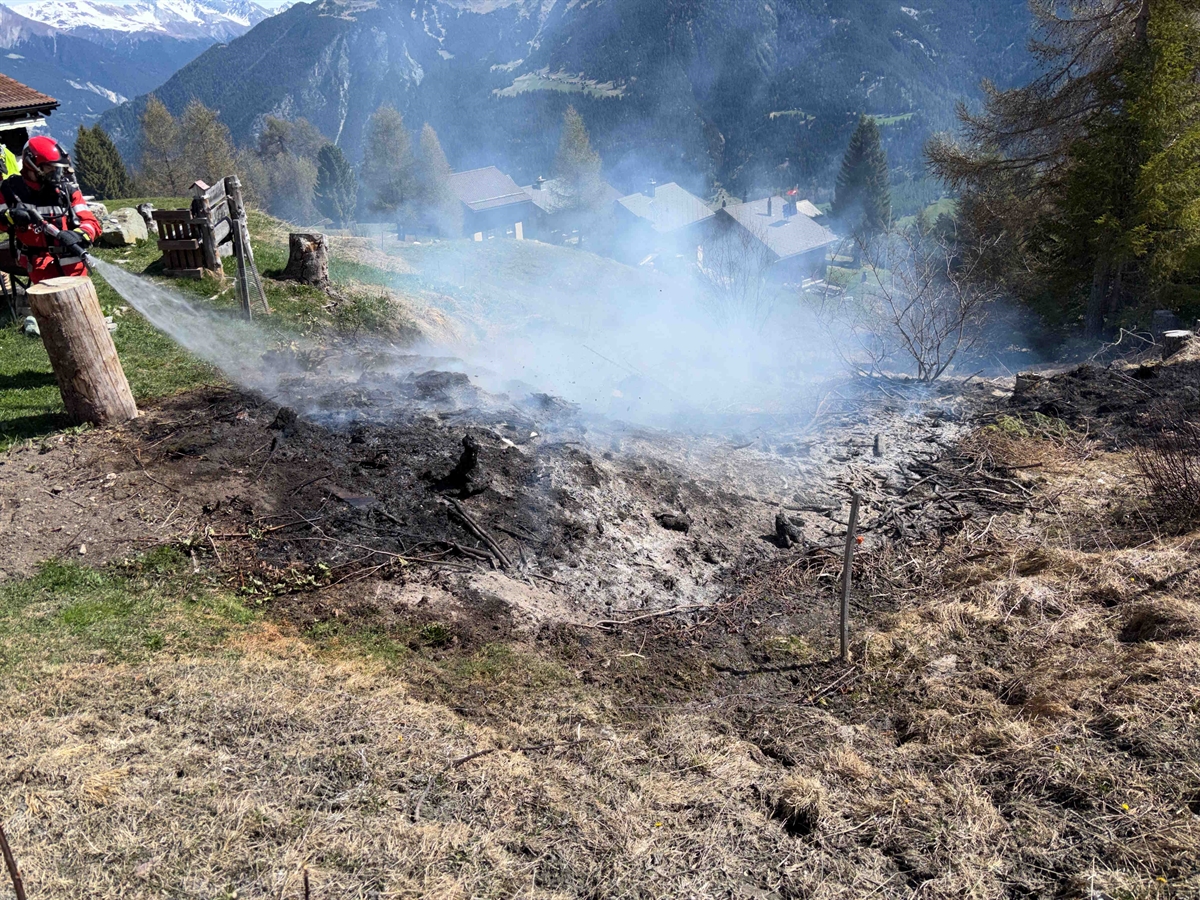 Übersichtsaufnahme mit Rauch ein Feuerwehrmann mit Wasserschlauch am löschen, verbrantes Wiesland nähe Waldgebiet und im Hintergrund Häuser und das Tal Surses.