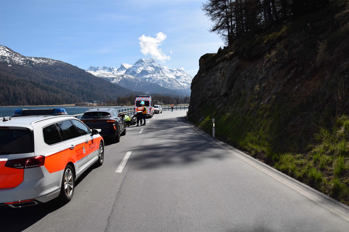 Übersichtsaufnahme der Malojastrasse, linksseitig im Hintergrund der Silsersee, Berge, blauer Himmel und rechtsseitig steile grüne Böschung und Felsen. Auf der linken Fahrbahn ein Patrouillenfahrzeug, zwei nicht am Unfallbeteiligte Pw`s, das verunfallte Motorrad samt Lenker, welcher durch das Ambulanzteam betreut wird und im Hintergrund die Ambulanz. 