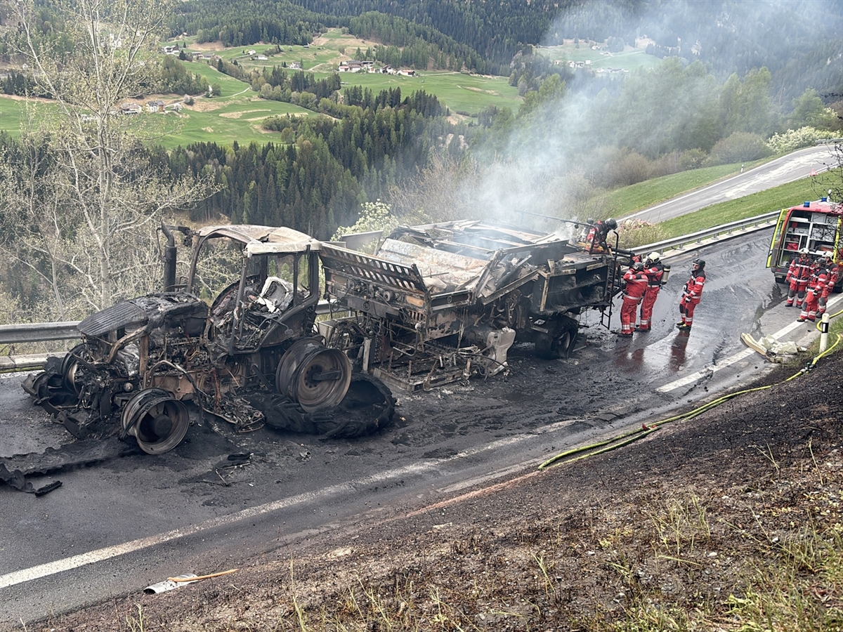 Die ausgebrannte Fahrzeugkombination auf der Strasse bergwärts in Richtung Ftan. Sieben Feuerwehrleute begutachten die rauchende Heupresse.