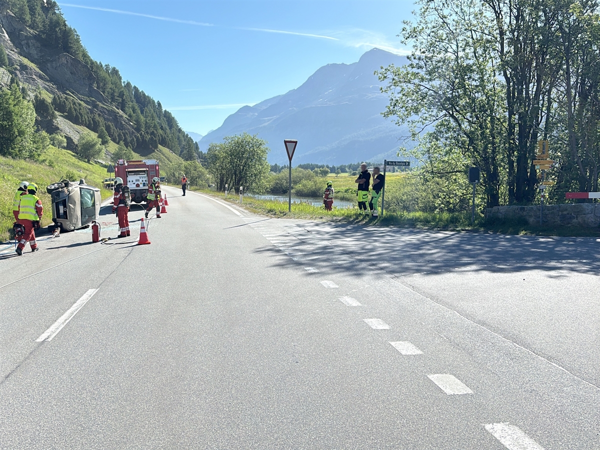 Der Kreuzungsbereich. Links am Strassenrand das total beschädigte Unfallauto. In dessen Bereich Einsatzkräfte. Im Hintergrund Berge welche sich vom heiteren Horizont abgrenzen.