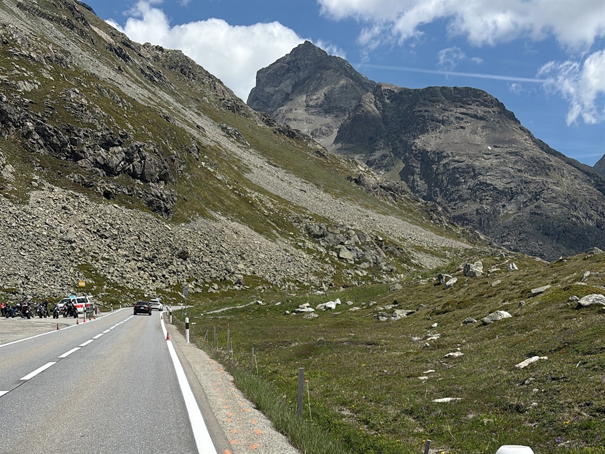 Rechts der Strasse auf dem Kiesbankett markierte Spuren. Angrenzend das steindurchsetzte Wiesland. Im Hintergrund Berge, die sich vom grösstenteils heiteren Horizont abgrenzen.