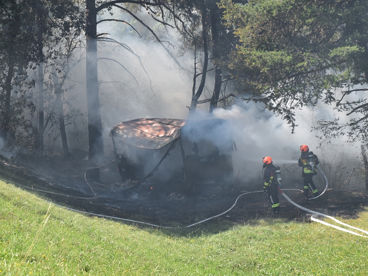 Zwischen Bäumen steht das rauchende Bienenhäuschen am Waldrand. Zwei Feuerwehrleute bekämpfen mit Wasser den Brandherd