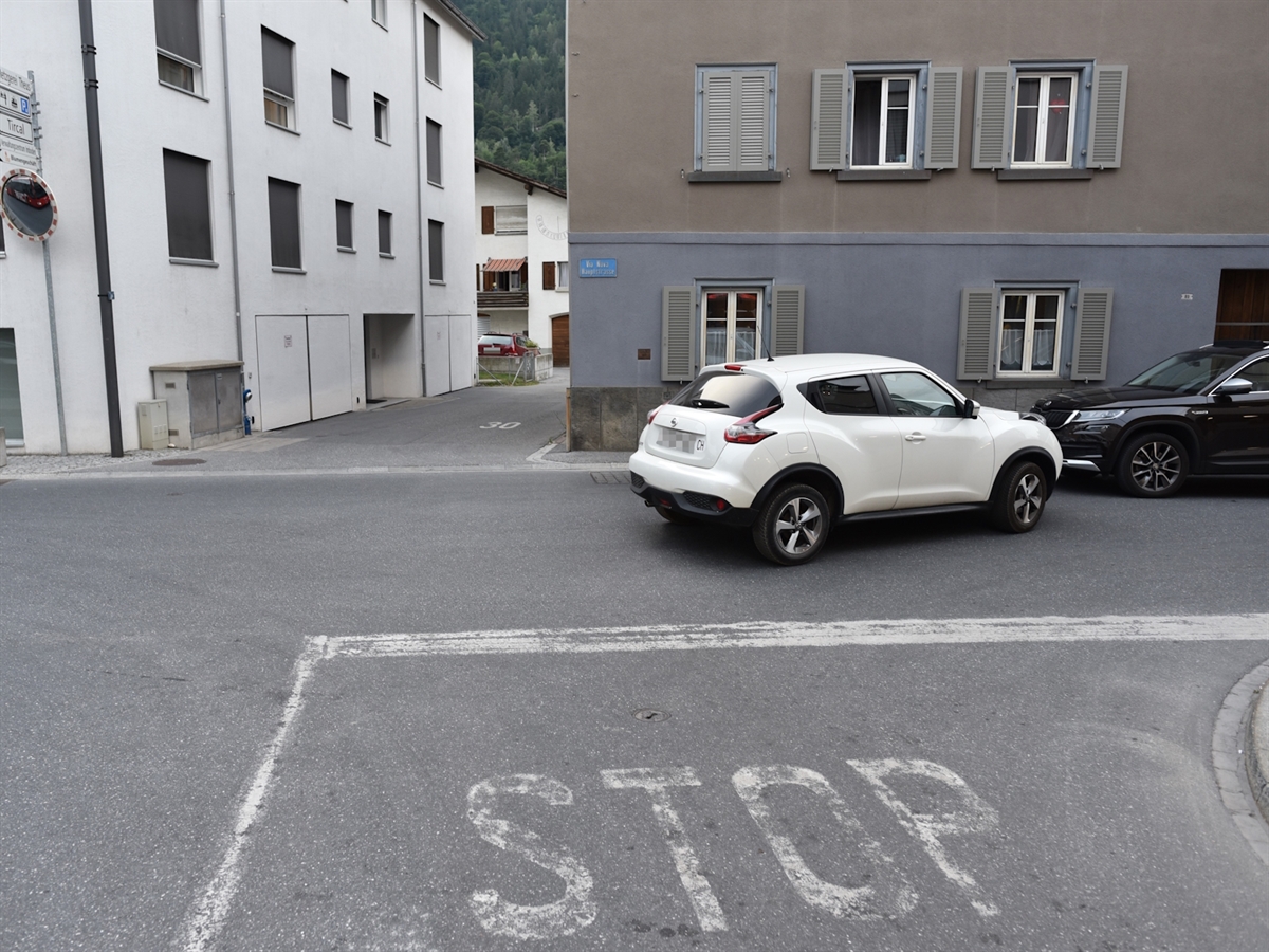 Auf der Via Nova stehen zwei Autos. Das weisse Auto der Frau steht mit Front gegen das schwarze Auto des Mannes. Auf der Fahrbahn ist gross ein *STOP* mit Haltebalken gezeichnet. Im Hintergrund steht eine Häuserreihe.