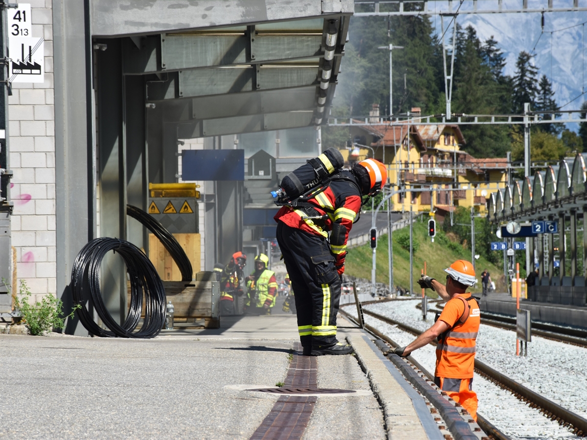 Ein Feuerwehrmann und ein Bahnangestellter im Gespräch beim Güterschuppen. Aus diesem quillt Rauch.