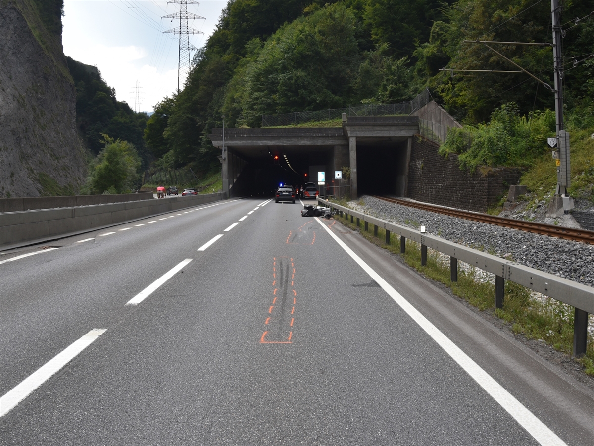 Das Ostportal des Chlustunnel mit der zweispurigen Fahrbahn. Auf dem rechten Fahrstreifen liegt unmittelbar vor dem Tunnel das verunfallte Motorrad, dessen Endlage und Spuren auf der Fahrbahn durch orange Markierungen gekennzeichnet sind. Im Tunnelbereich stehen Einsatzfahrzeuge mit eingeschaltetem Blaulicht, während rechts neben der Strasse eine Bahnlinie verläuft.