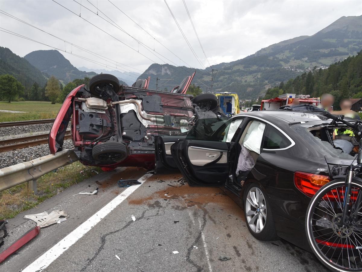 Ein rotes Fahrzeug liegt auf der Seite und ist mit der Leitplanke verkeilt. Davor steht das zweite Unfallfahrzeug, ein schwarzes Auto. Im Hintergrund sind Einsatzfahrzeuge sowie die Bahnlinie zu sehen.