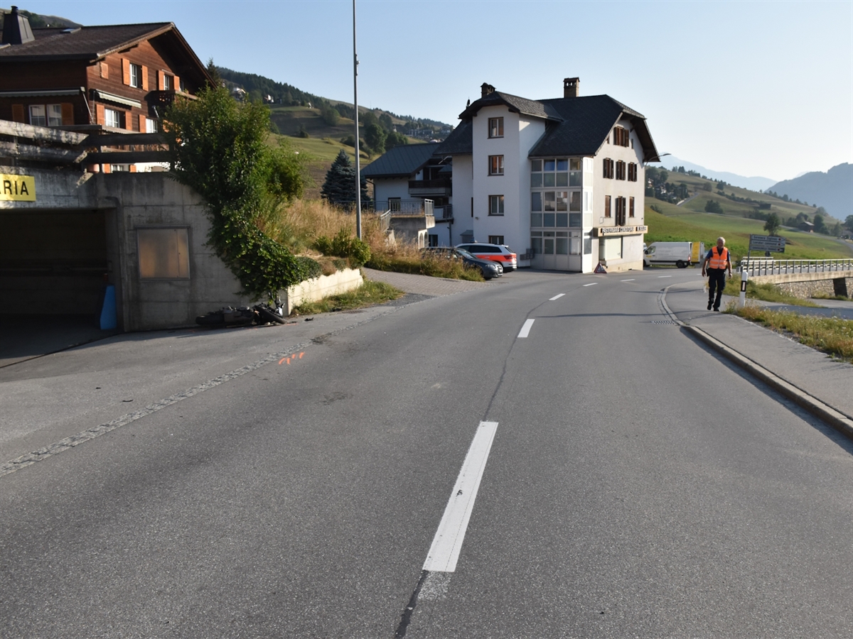 Die Unfallörtlichkeit mit dem liegenden Motorrad vor einer Mauer bzw. Garagenzufahrt. Auf dem Trottoir steht ein Polizist. Im Hintergrund auf einem Parkplatz steht ein Patrouillenfahrzeug. Häuser säumen den linken Strassenrand.