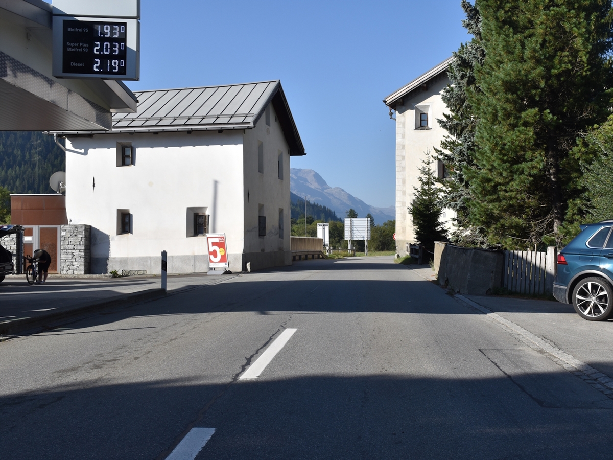 Links die Tankstelle. Die Hauptstrasse wird zwischen zwei Häusern verengt. Im Hintergrund Bergpanorama und wolkenloser Himmel.