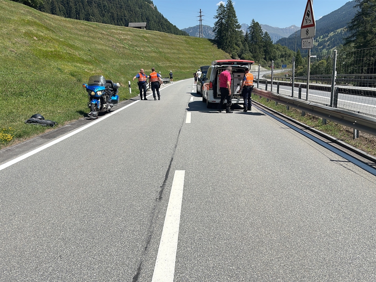 Rechts der Hauptstrasse die Nationalstrasse. Auf der Hauptstrasse: Ein Polizeiauto, das beteiligte Motorrad und Polizisten. Im Hintergrund Bergpanorama und wolkenloser Himmel.