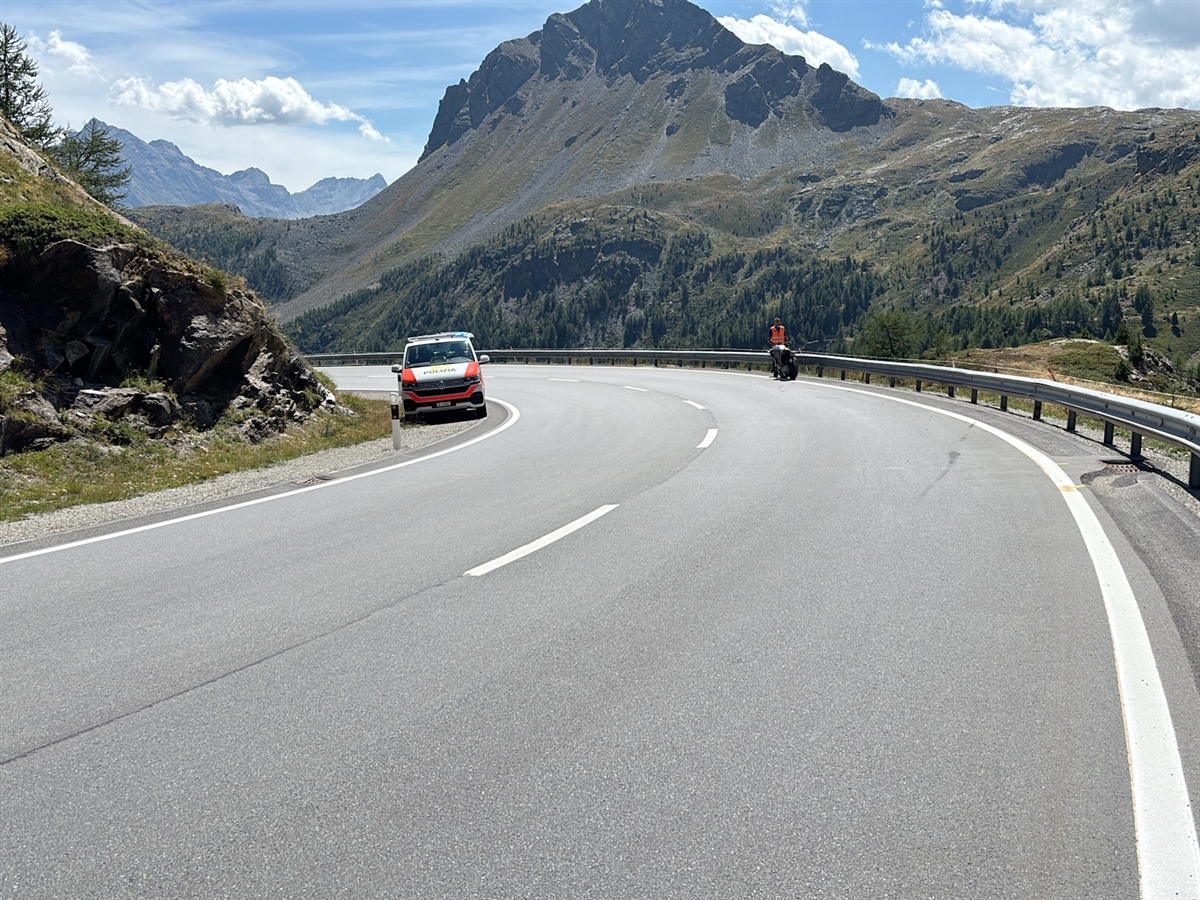Die Linkskurve der Berninastrasse. Am rechten Strassenrand steht ein Polizist hinter dem auf dem Seitenständer abgestützten Unfallmotorrad. Links an der Fahrbahn steht ein Polizeiauto. Auf der talwärtsführenden Fahrbahn sind Kratzspuren ersichtlich.