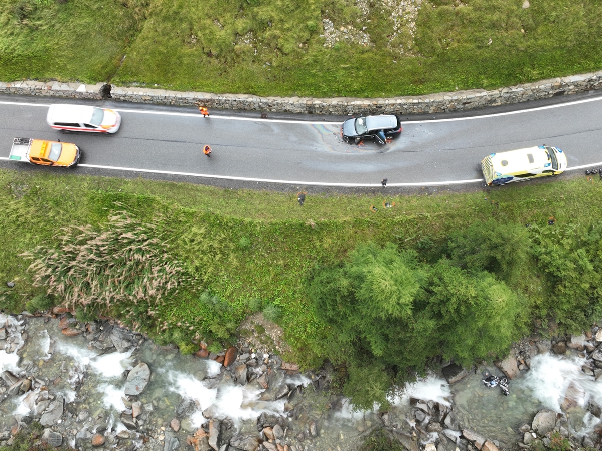 Drohnenaufnahme der Rechtskurve, in welcher das Auto ganz am rechten Strassenrand steht. Ein Polizeiauto, ein Rettungswagen sowie ein Fahrzeug des Tiefbauamtes säumen die Strasse weiter. Am unteren Bildrand liegt das Motorrad im Bach.
