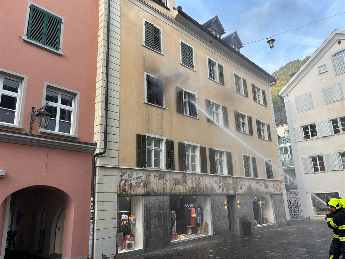 Das vierstöckige Mehrfamilienhaus in der eng umbauten Churer Altstadt. Ein Feuerwehrmann bekämpft die Flammen im zweiten Obergeschoss mit Wasser durch ein Fenster. Aus einem weiteren Fenster steigt Rauch hervor.
