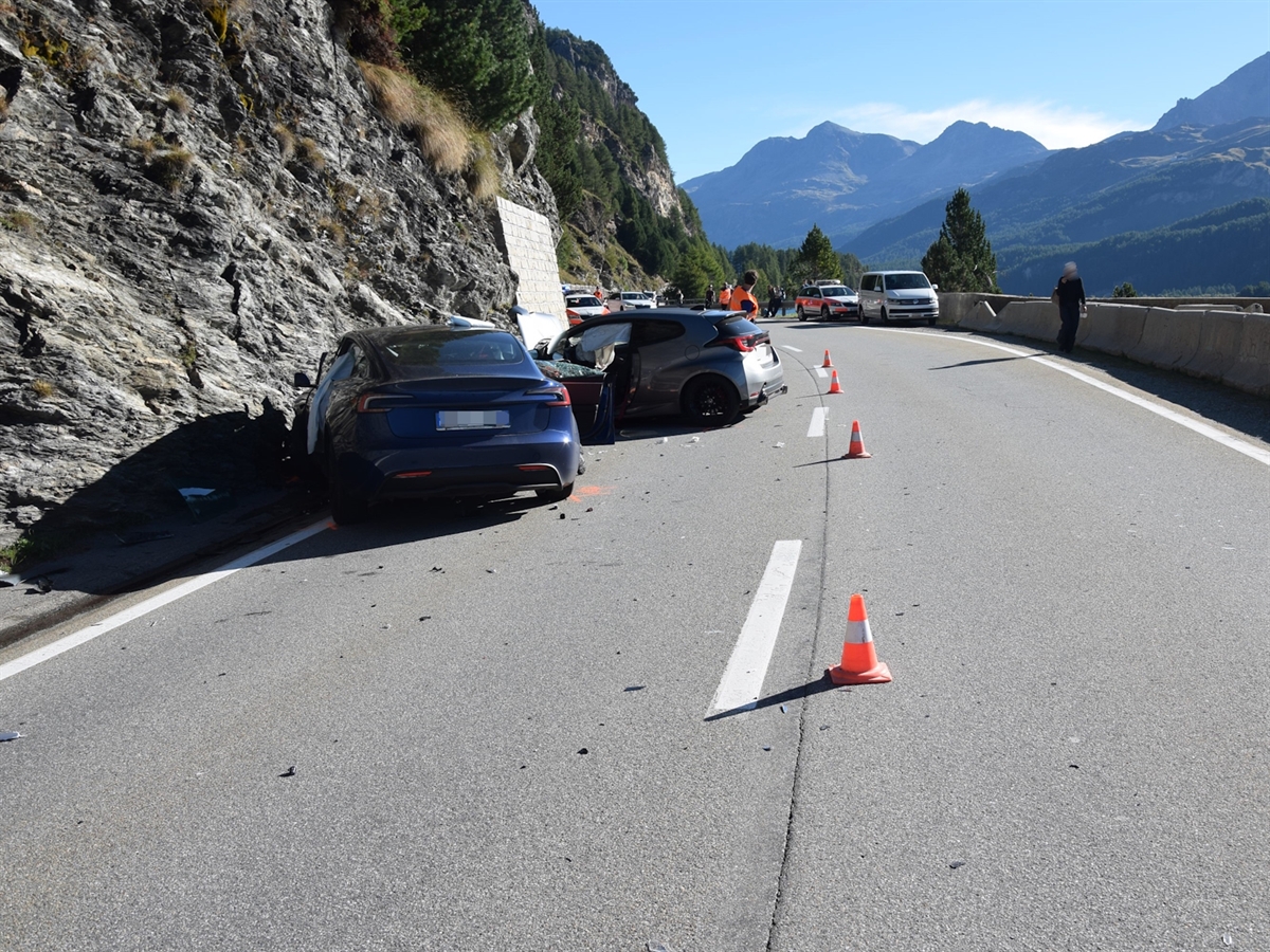 Die beiden Unfallautos stehen abgedreht gegen eine Felswand auf der Fahrspur der Schweizerin. Im Hintergrund Einsatzkräfte, Einsatzfahrzeuge sowie Berge, die sich vom blauen Horizont abgrenzen.