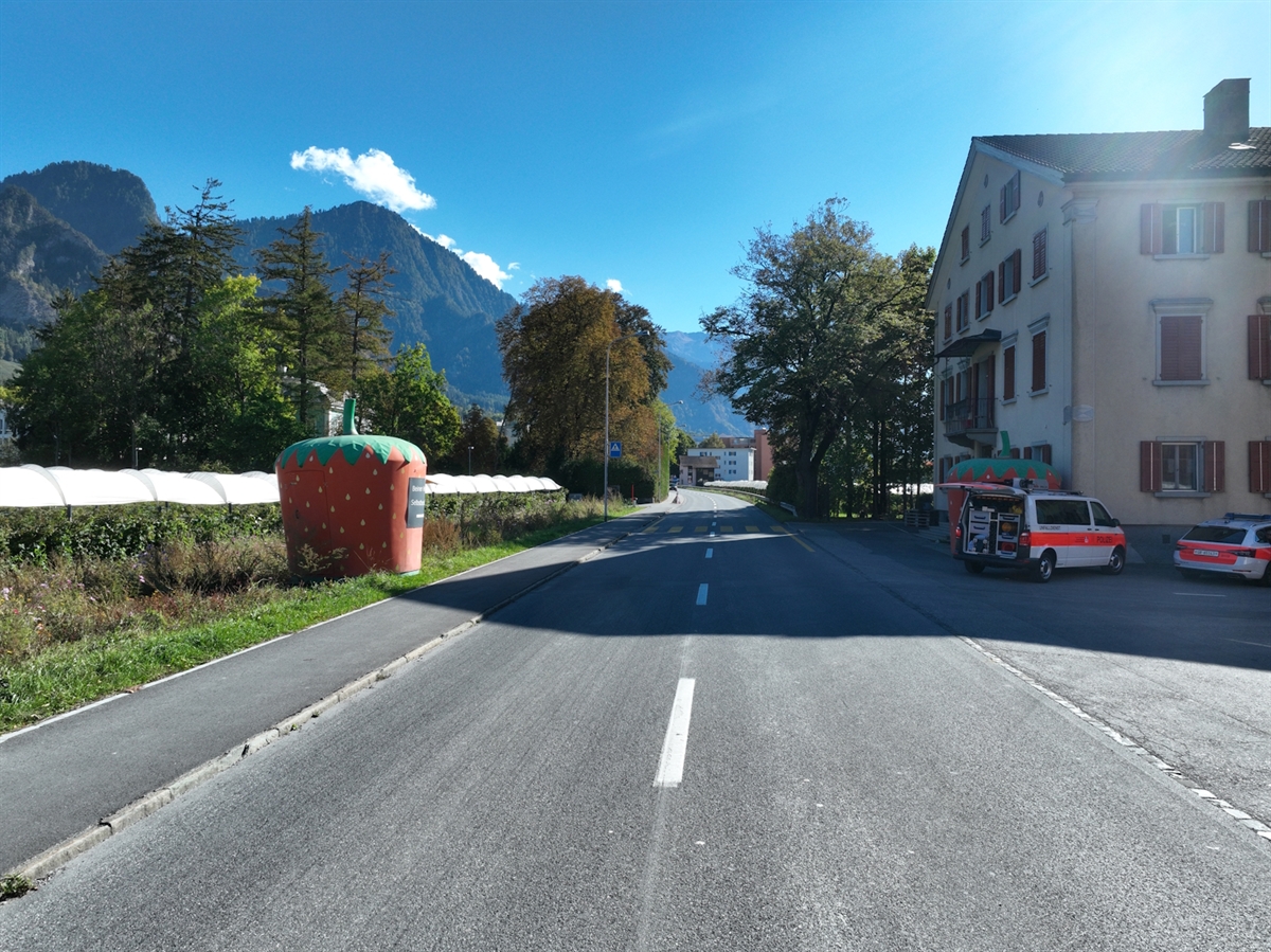 Die Unfallörtlichkeit beim Neuhof in Landquart. Der Fussgängerstreifen ist in der Bildmitte im Schatten erkennbar. Rechts wird die H13 durch den Vorplatz des Neuhofs und links durch ein Trottoir begrenzt. Zwei Patrouillenfahrzeuge sind auf dem Vorplatz beim Haus Neuhof abgestellt.