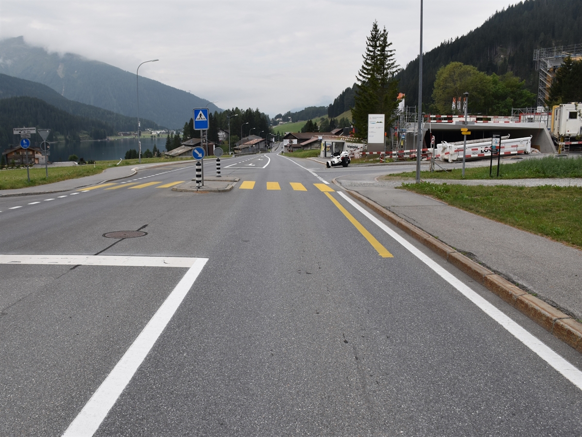 Die Prättigauerstrasse mit einer Mittelinsel und einem Fussgängerstreifen im Bereich der Einfahrt zur Höhenklinik. Rechts befindet sich eine Baustelle, links sind der Davoser See sowie die umliegende Berglandschaft zu sehen.
