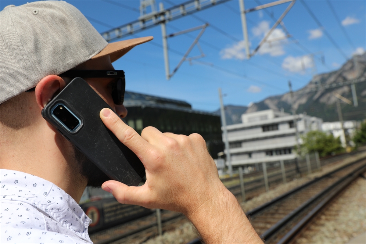 Eine zweifelhafte Gestalt mit Cap und Sonnenbrille telefoniert mit dem Mobiltelefon. Im Hintergrund Bahngeleise, Gebäude, Berglandschaft mit blauem Himmel.