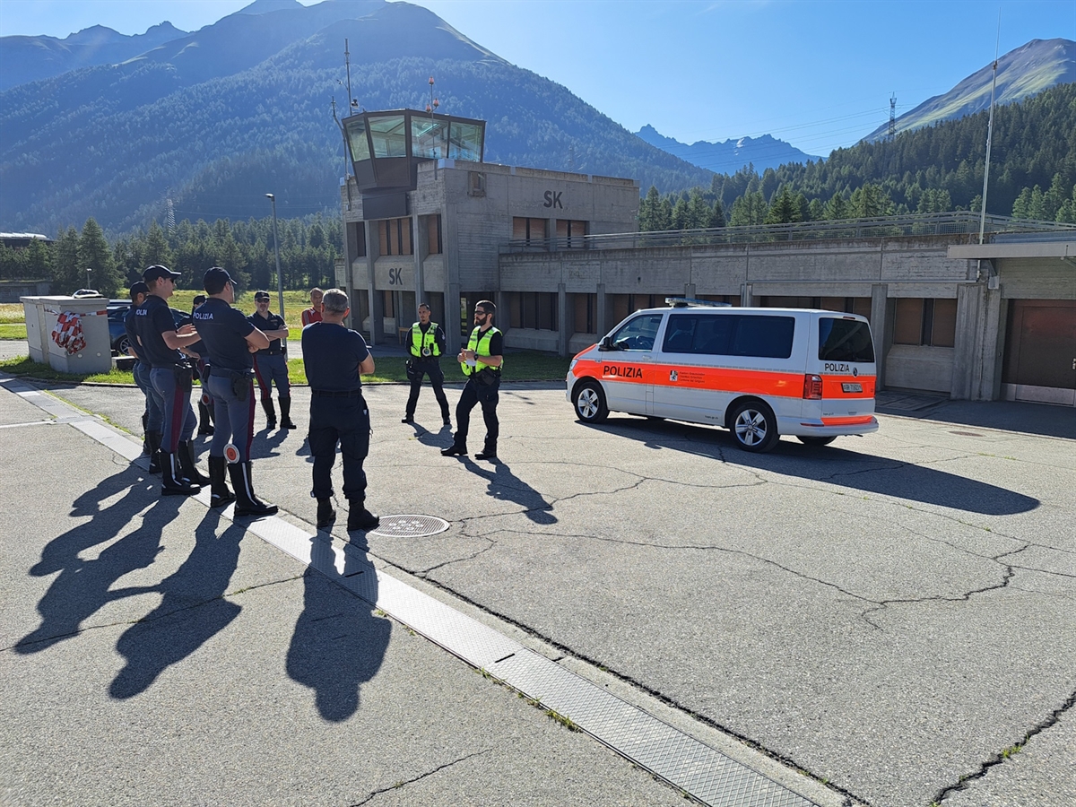 Zwei Instruktoren stehen in einer Bergkulisse vor einem Polizeiauto. Ihnen gegenüber weitere Einsatzrkfäte. 