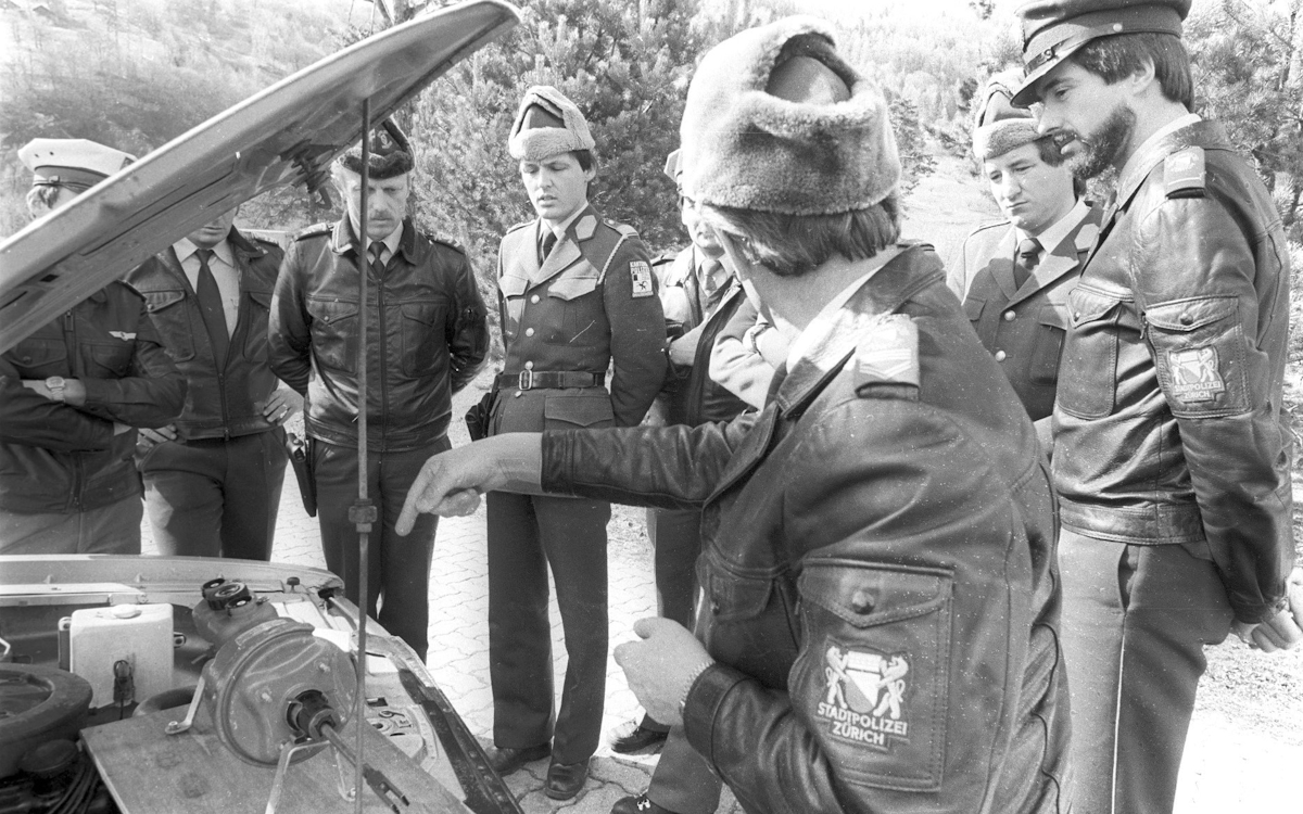 Schwarz-weiss-Foto aus den späten 70er-Jahren mit acht Polizisten beim Begutachten eines Bremsmodells. Als Präsentiertisch wurde der Motorraum eines Autos verwendet.