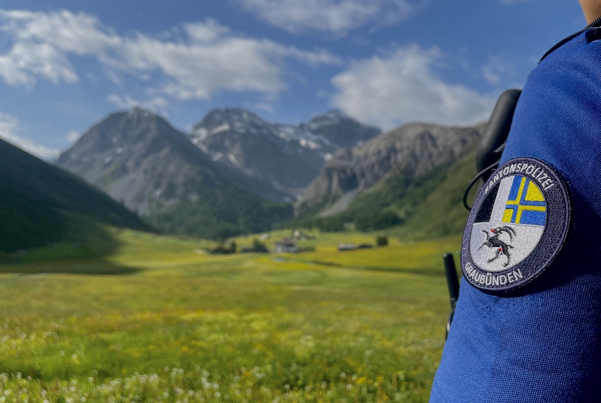 Blick über die Schulter eines Polizisten mit Armbatch ins Tal mit Wiese und Bergen