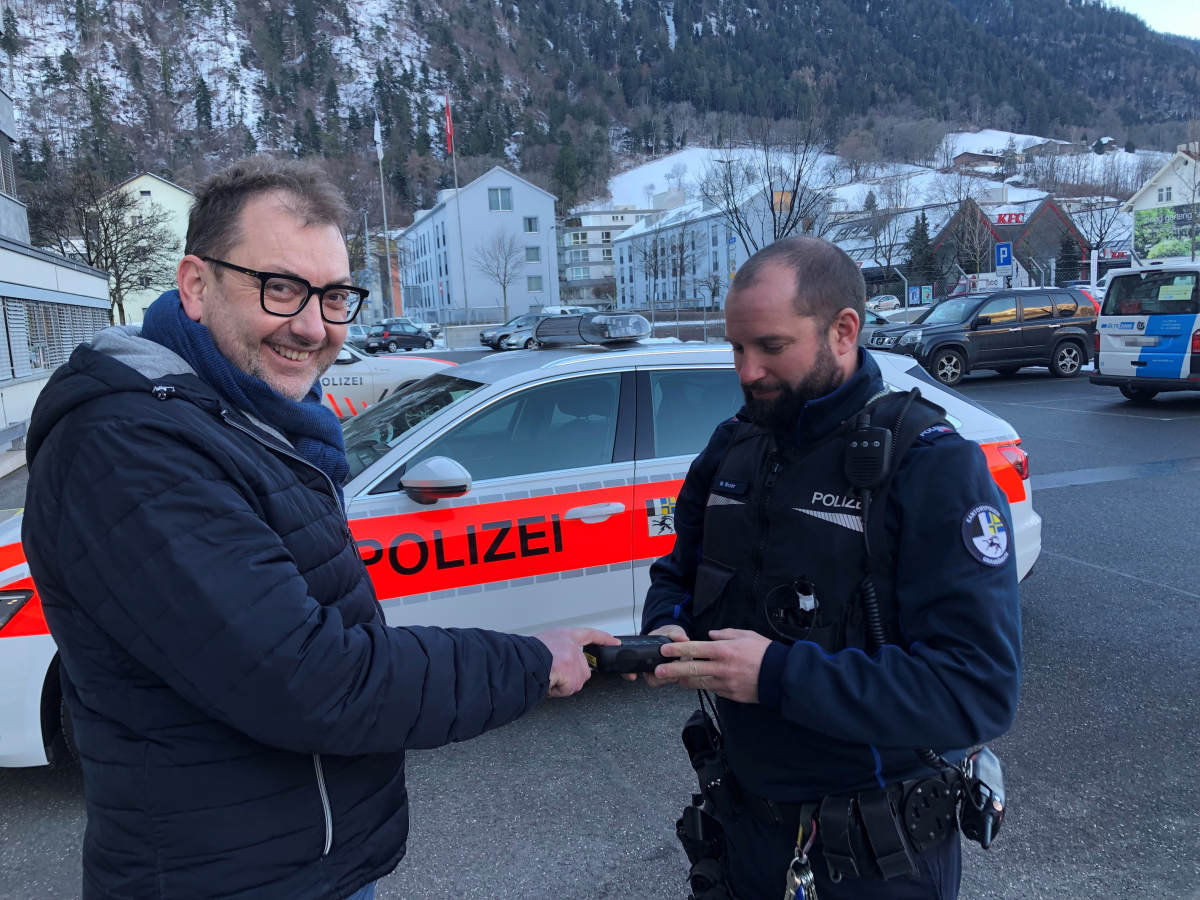 Regierungsrat Peter Peyer steht vor einem Patrouillenfahrzeug der Kantonspolizei Graubünden und lässt sich von einem Polizisten seine Fingerabdrücke scannen.