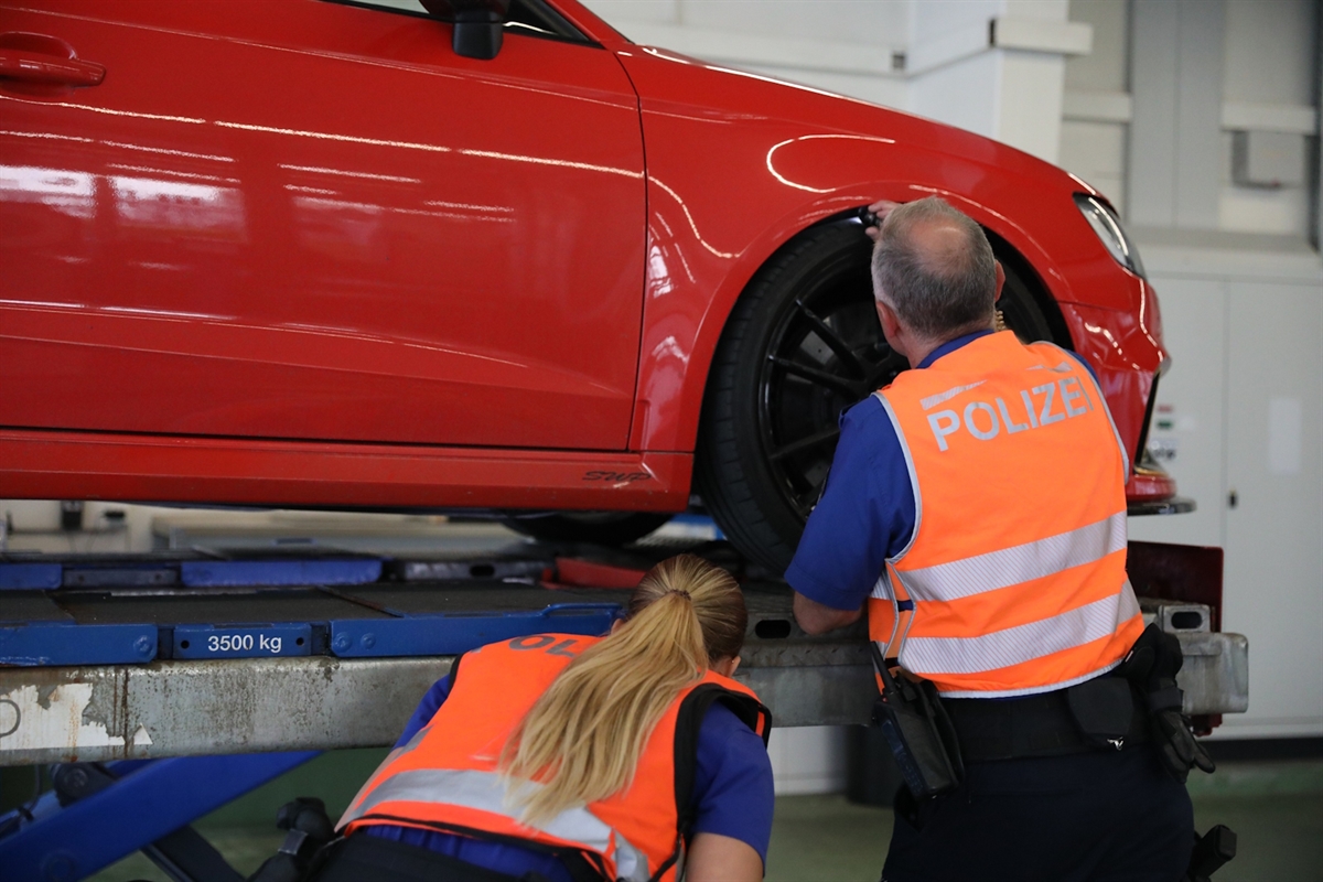 Eine Polizistin und ein Polizist bei der technischen Kontrolle eines Autos, welches angehoben auf einem Lift steht. 