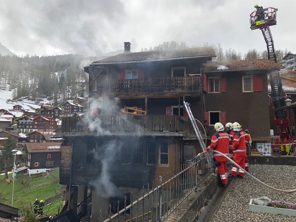 Das Holzhaus, daraus quillt Rauch hervor. Auf einer Terrasse stehen Feuerwehrleute, die das Gebäude sichern. Auf einer Drehleiter ein weiterer Feuerwehrmann.