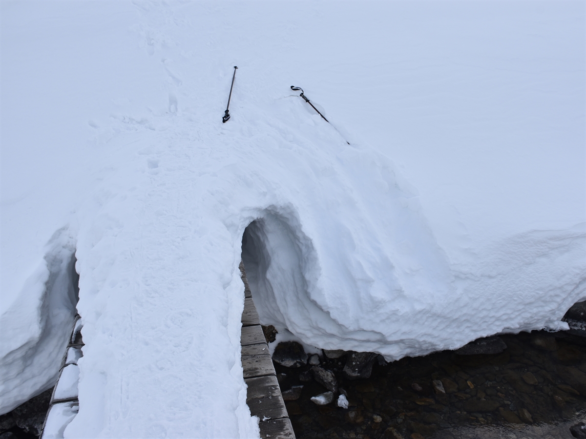 Die schneebedeckte Brücke und der angrenzende Steilhang. Rechts sind Rutschspuren in den Bach sichtbar. Im Schnee liegen zwei Tourenstöcke