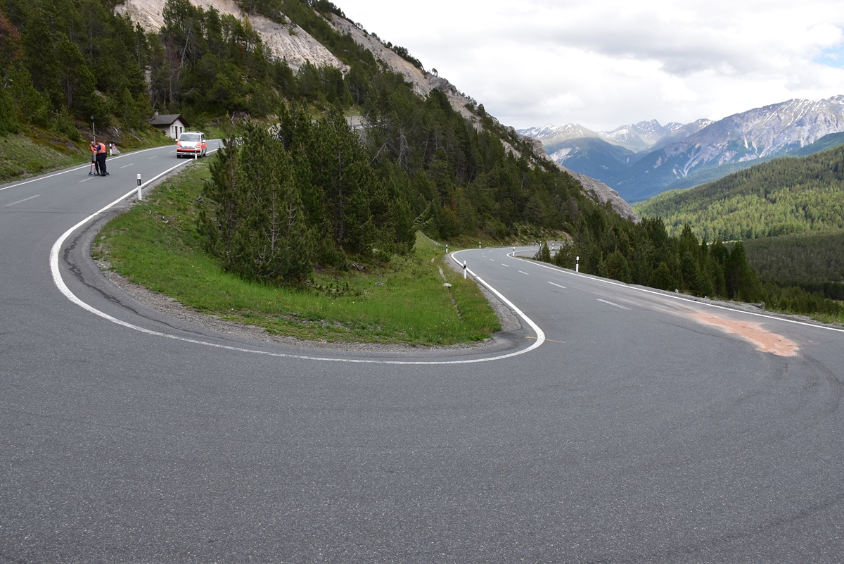 Von links nach rechts: Die von oben nach unten führende Strasse. Im Vordergrund die Linkskurve, links oben zwei Polizisten, rechts nach der Wendekehre die Oelspur, auf welcher Oelbinder liegt.