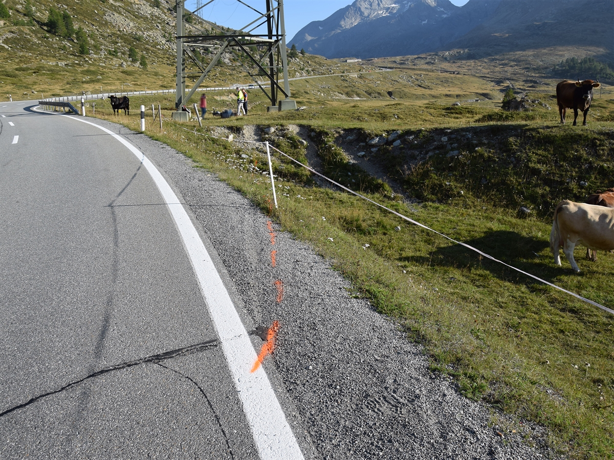 Linkskurve der Berninastrasse in Richtung Hospiz gesehen. Am rechten Strassenrand sind Pneuabdruckspuren im Kies sichtbar. Im Hintergrund der Strommast mit dem gestürzten Motorradlenker sowie Einsatzkräfte. Mehrere Kühe weiden auf der Alpwiese