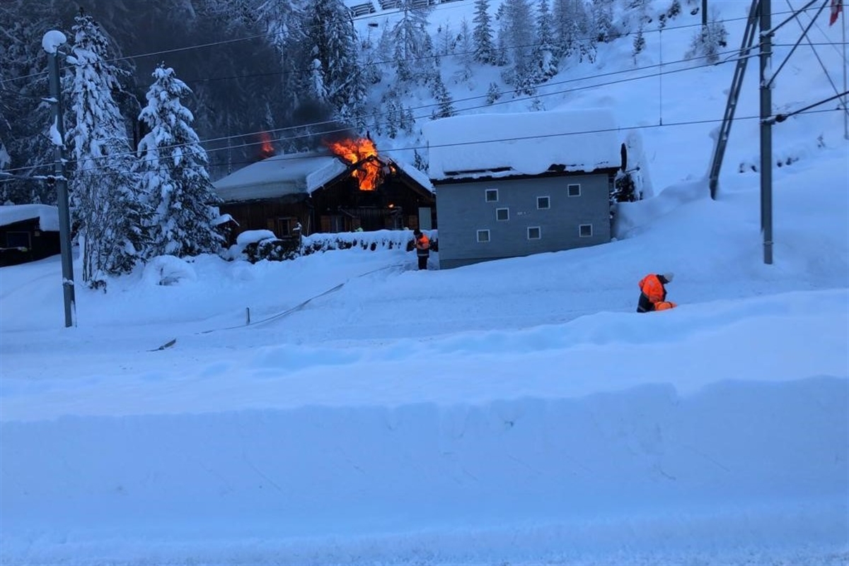 Winterlandschaft mit dem Ferienhaus bei Punt Ota in Bergün. Aus dem Dachgiebel treten Flammen heraus. Im Vordergrund die Fahrleitung der RhB.