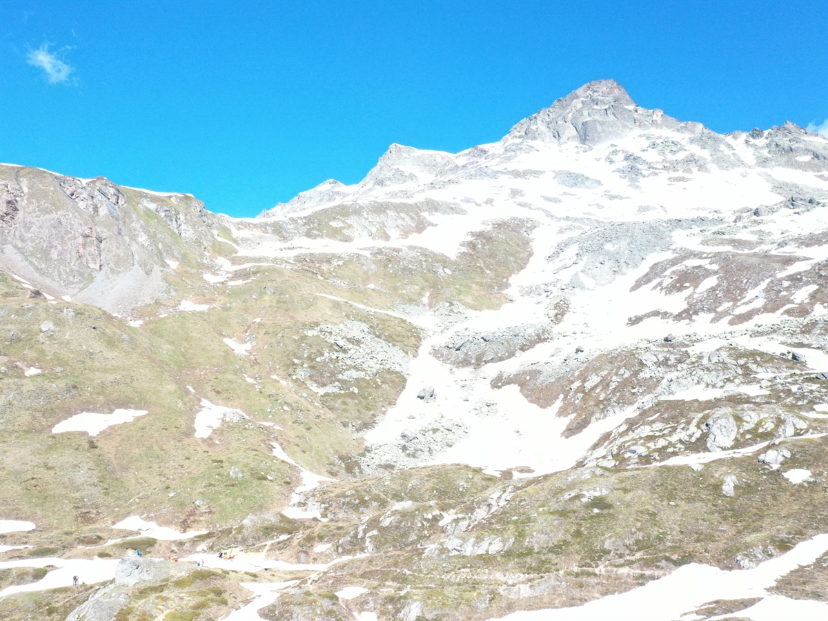 Geländeaufnahme mit Wiesland und Schneefeldern. Als Begrenzung zum Horizont (blauer Himmel) der Piz Neir.