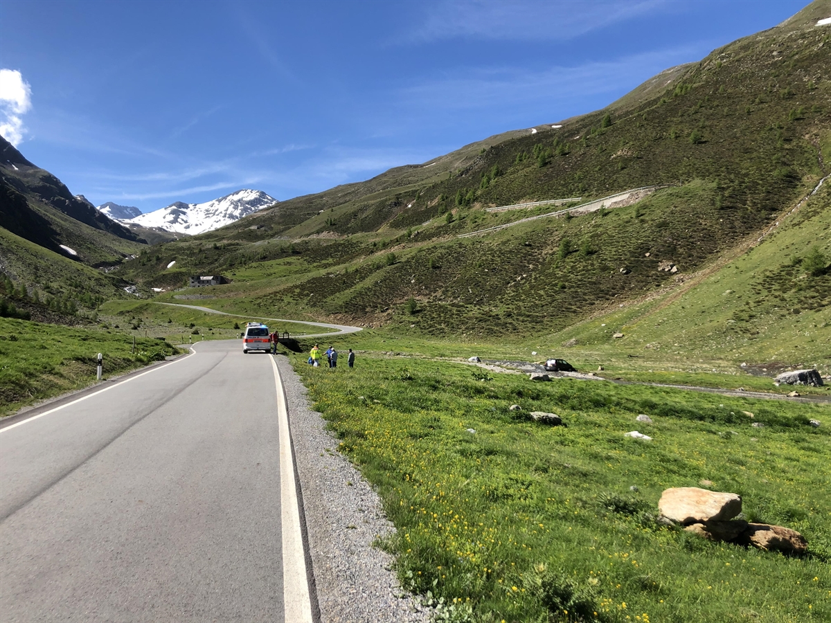 Blick in Richtung Umbrailpass. Rechtsseitig ist das Wiesland und ein Bach. Im Bach ist das Auto zu sehen. Es steht ein Polizeibus auf der rechten Strassenseite und daneben sind die beteiligten Personen inkl. Rettungkräfte.