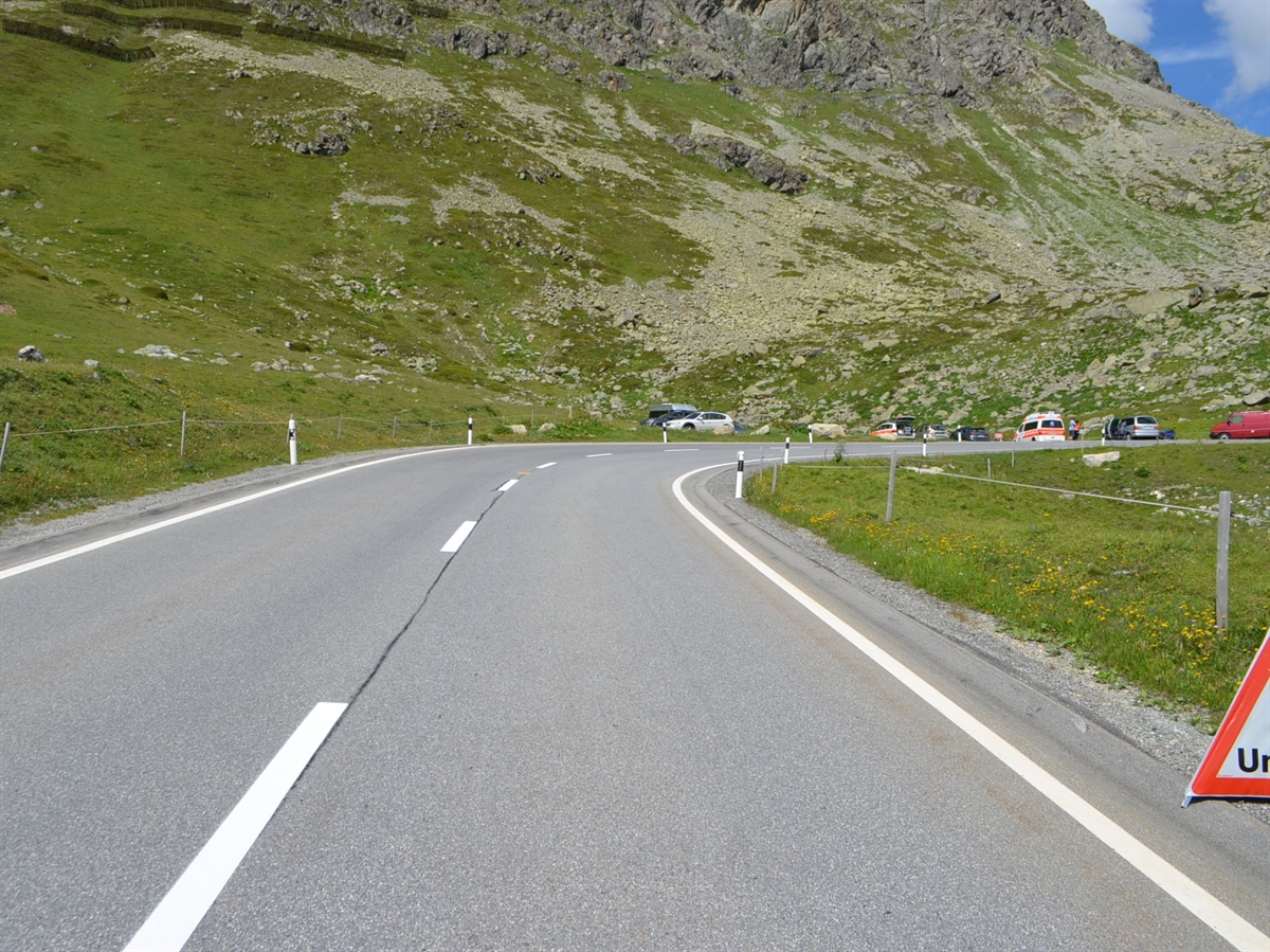 Das Ende der langen Geraden mit der langgezogenen Rechtskurve. Im Hintergrund der Parkplatz linksseitig der Kurve mit einem Patrouillenfahrzeug, einer Ambulanz sowie dem verunfallten Töff und dem Auto.